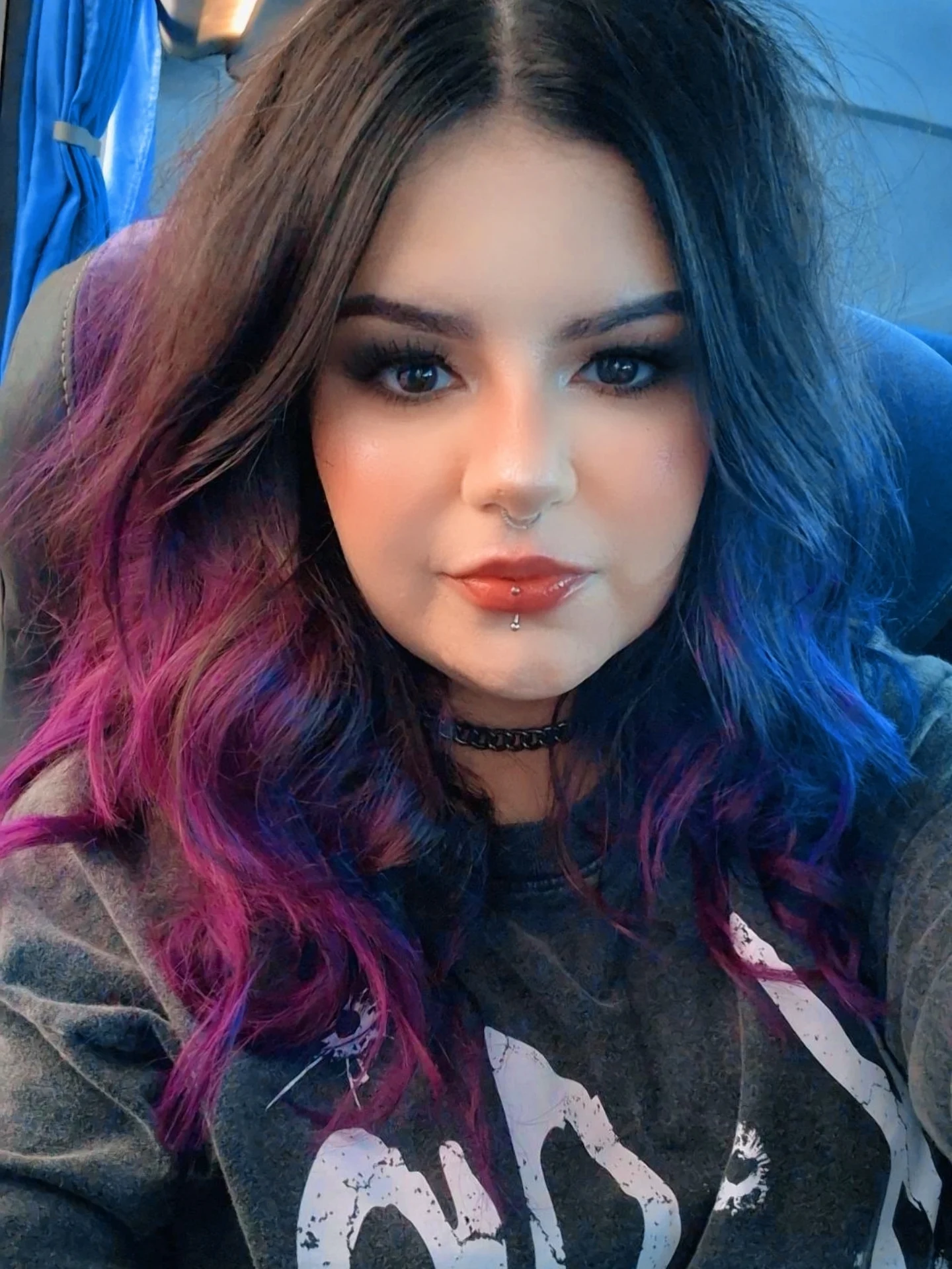 Mulher de cabelo ondulado médio preto com pontas roxas, pele branca, olhos castanho-escuros. Ela usa maquiagem escura, gloss vermelho, um piercing no septo nasal e um piercing no lábio inferior. Ela veste uma camiseta cinza estampada e uma gargantilha prateada. O fundo mostra o interior desfocado de um ônibus.
